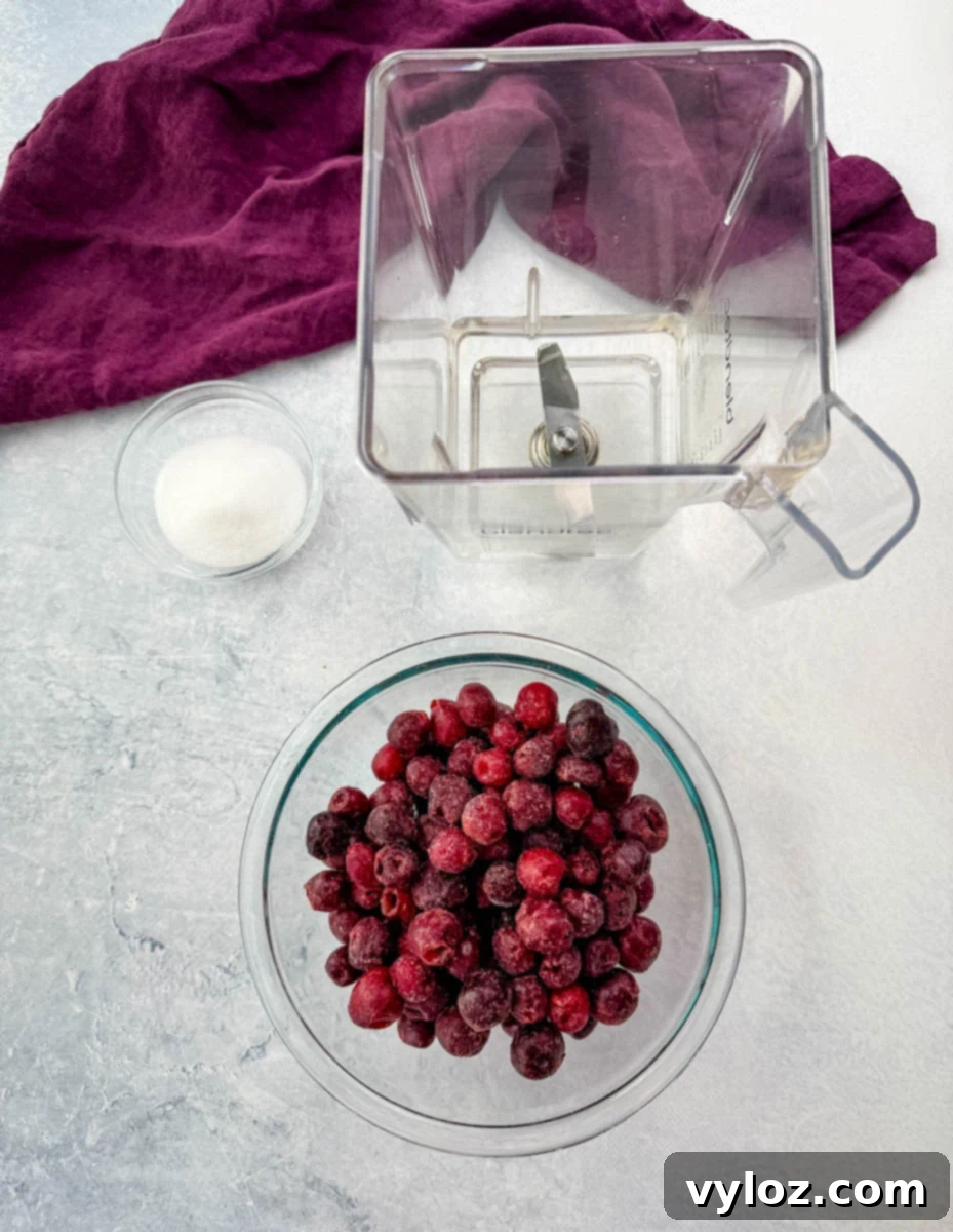 A flat-lay image displaying a clear blender jar, a bowl of frozen cherries, and a small glass bowl of sugar, ready to be blended into a cherry slushi. The background has a neutral textured surface with a deep purple cloth.