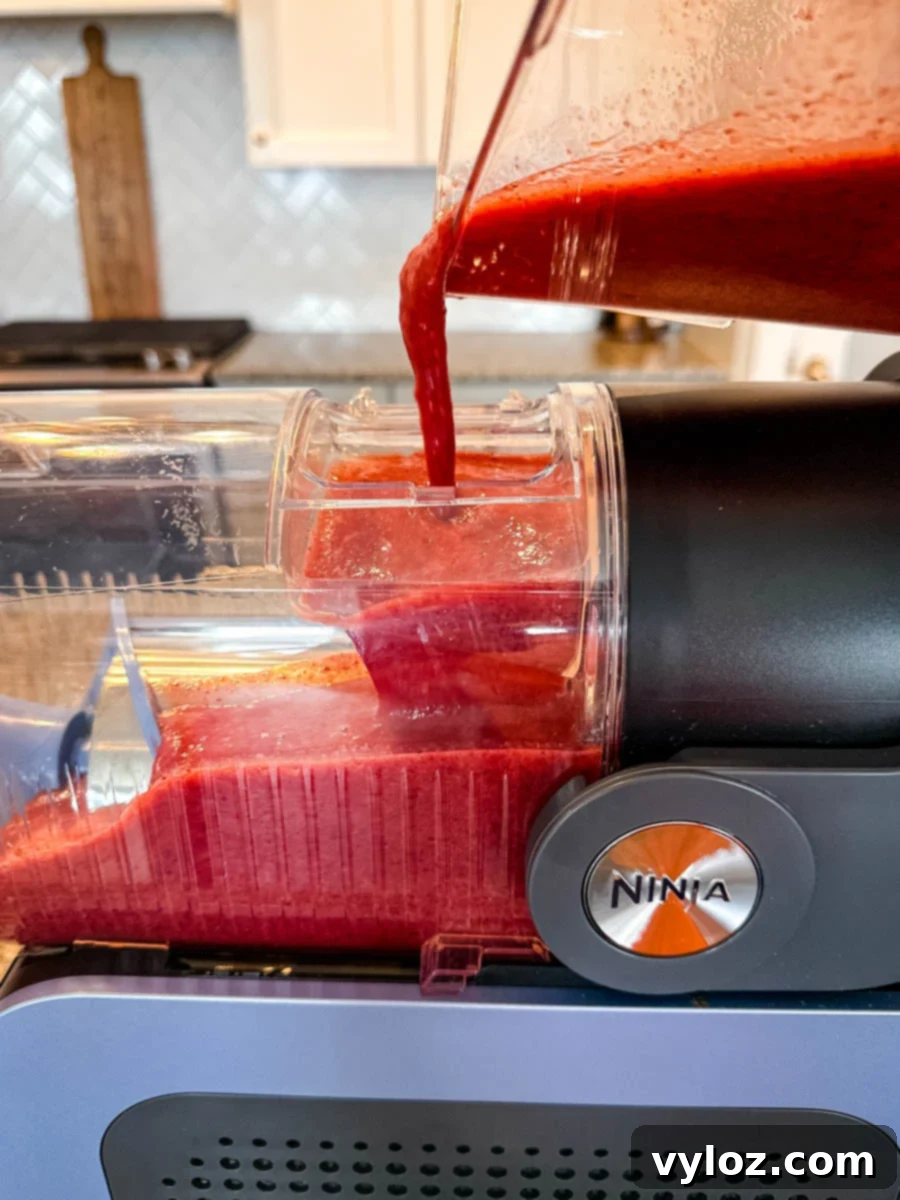 A close-up of a vibrant red cherry mixture being poured from a blender into the Ninja Slushi Machine. The background features a bright kitchen with a white backsplash and wooden cutting board.
