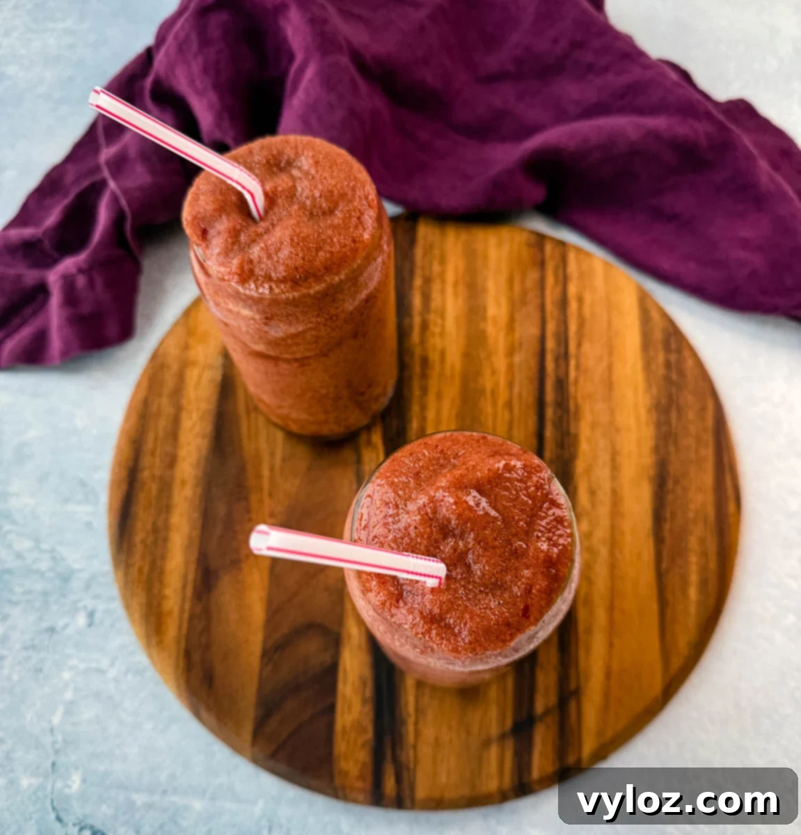 An overhead view of two tall glasses filled with frozen cherry slushies, placed on a wooden serving board. The drinks are garnished with striped straws, and a purple cloth adds contrast in the background.