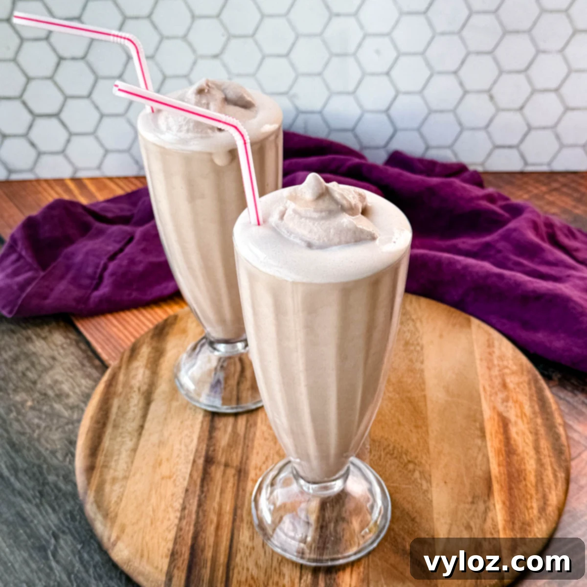 Two glasses of homemade Wendy’s Frosty-style milkshakes served with pink-striped straws, placed on a wooden board with a purple cloth in the background and a hexagonal tile backsplash.