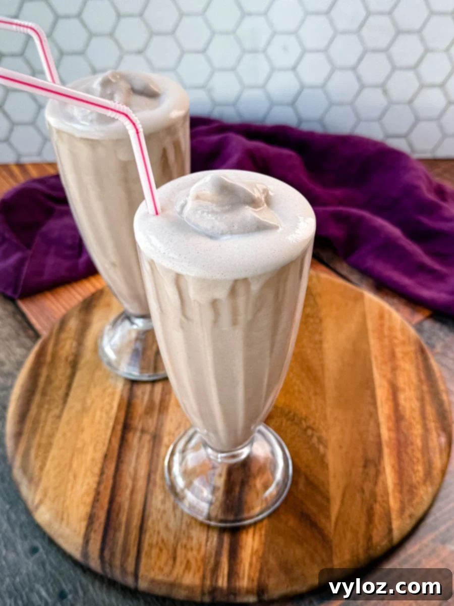 Two glasses of homemade Wendy’s Frosty-style milkshakes served with pink-striped straws, placed on a wooden board with a purple cloth in the background and a hexagonal tile backsplash.