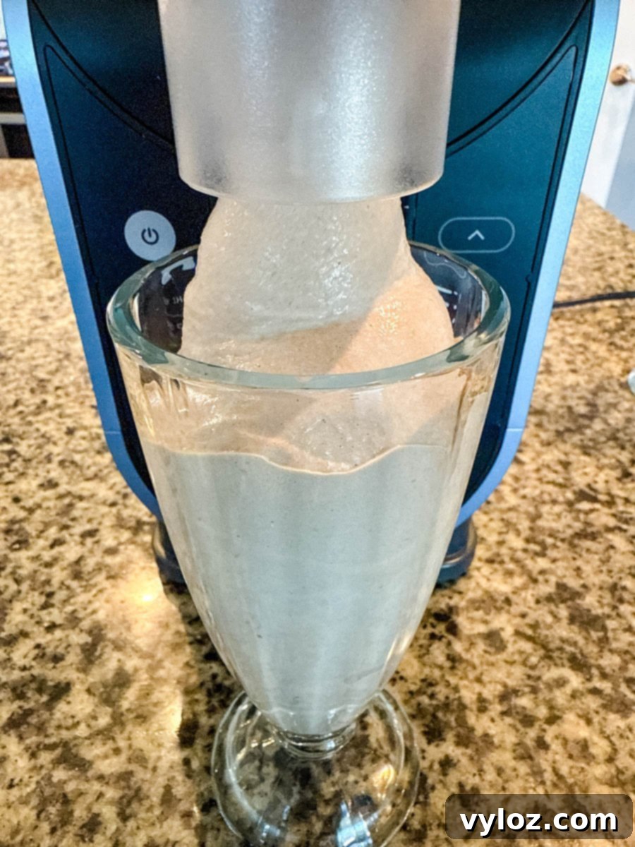 A thick, creamy Wendy’s Frosty-style milkshake being dispensed from a Ninja slushie machine into a tall glass on a granite countertop.