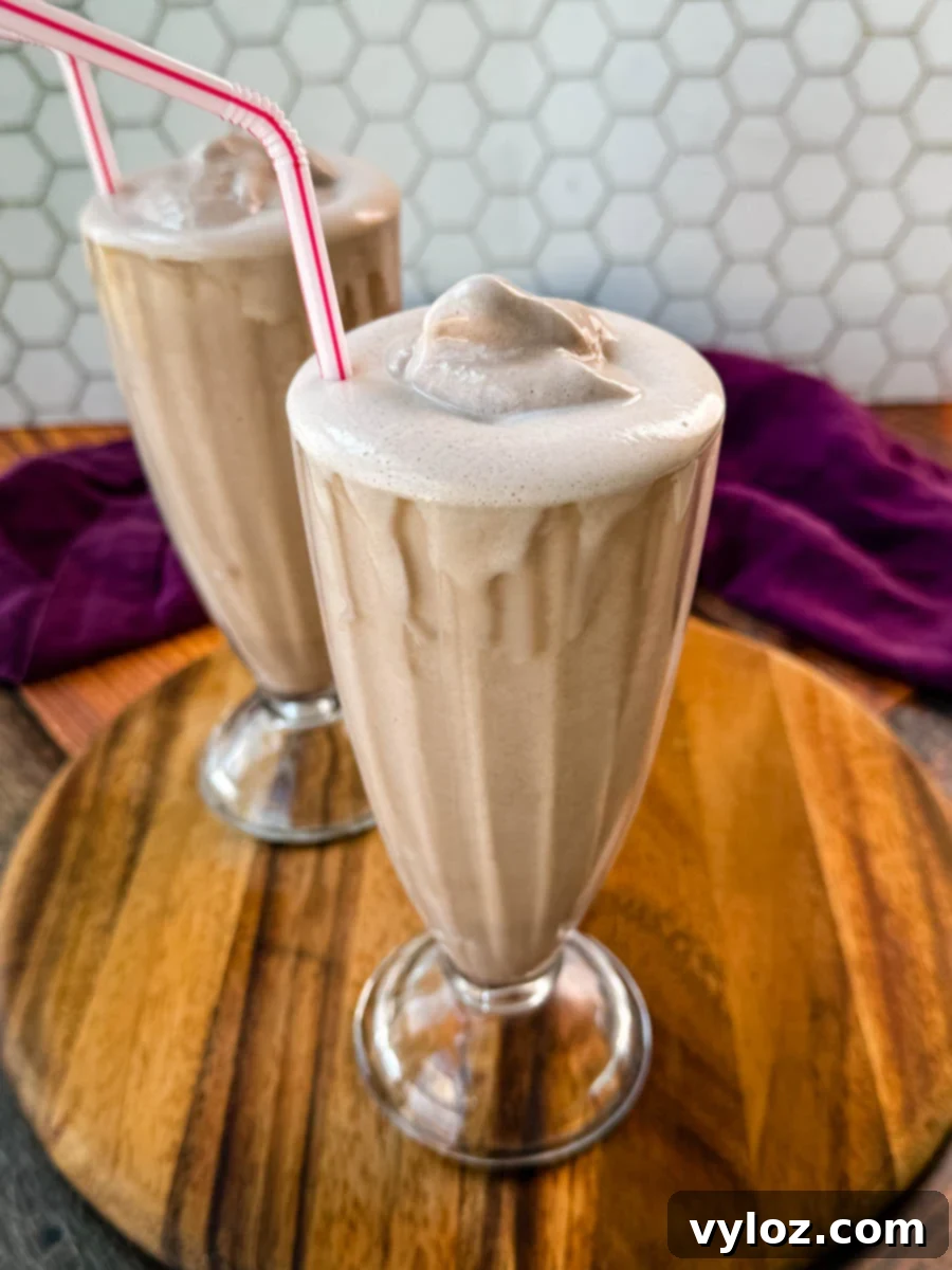 Two glasses of homemade Wendy’s Frosty-style milkshakes served with pink-striped straws, placed on a wooden board with a purple cloth in the background and a hexagonal tile backsplash.