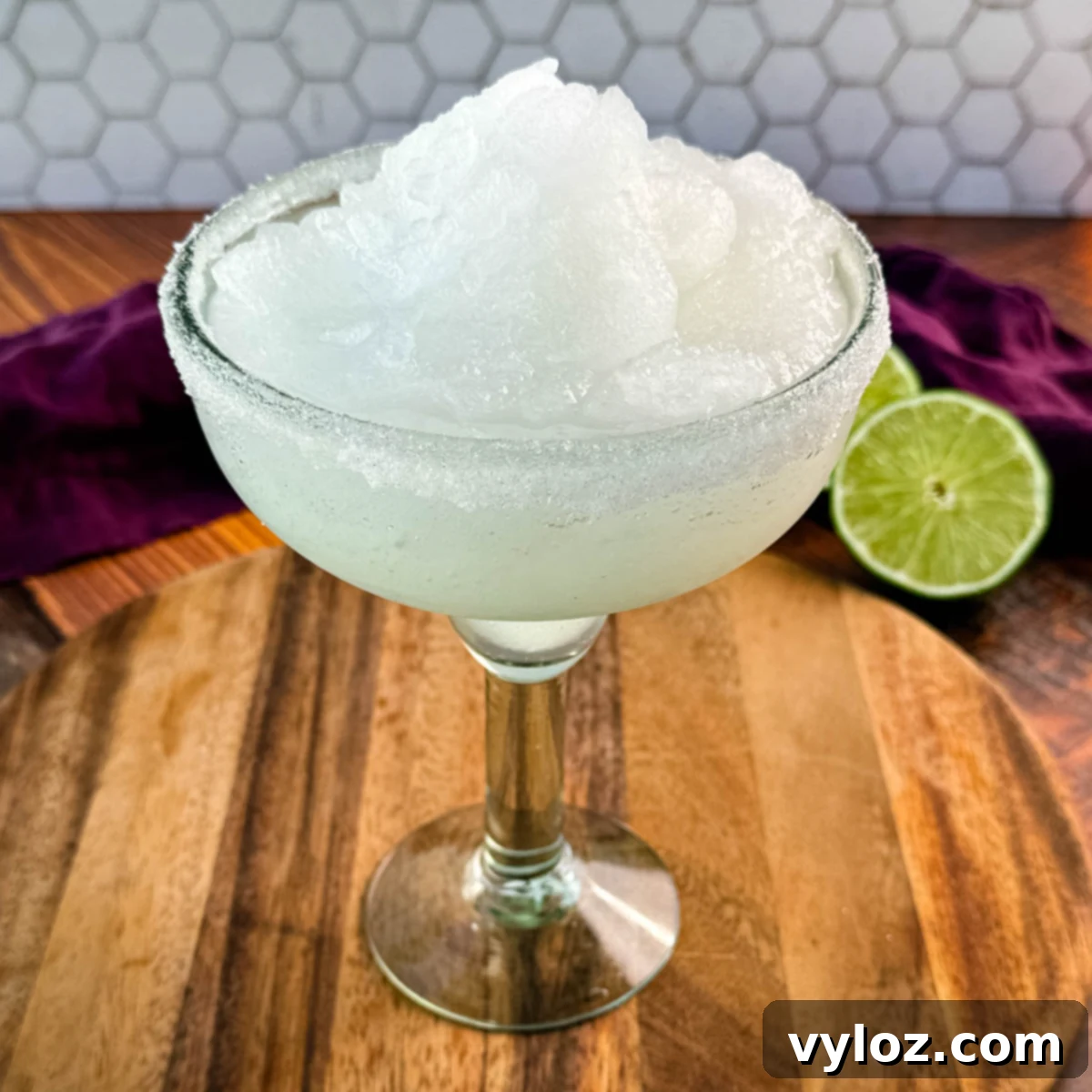 A close-up of a margarita glass filled with frozen slush, highlighting the fluffy, snow-like texture of the drink. The sugar-rimmed glass sits on a rustic wooden board.