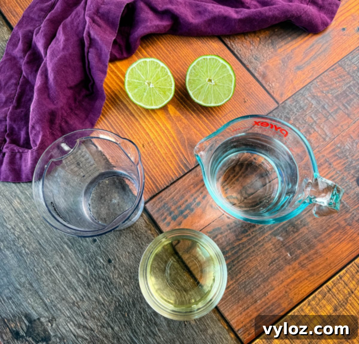 A flat lay of margarita ingredients including a measuring cup of water, lime halves, and small bowls of simple syrup and tequila, displayed on a wooden table with a dark purple napkin.