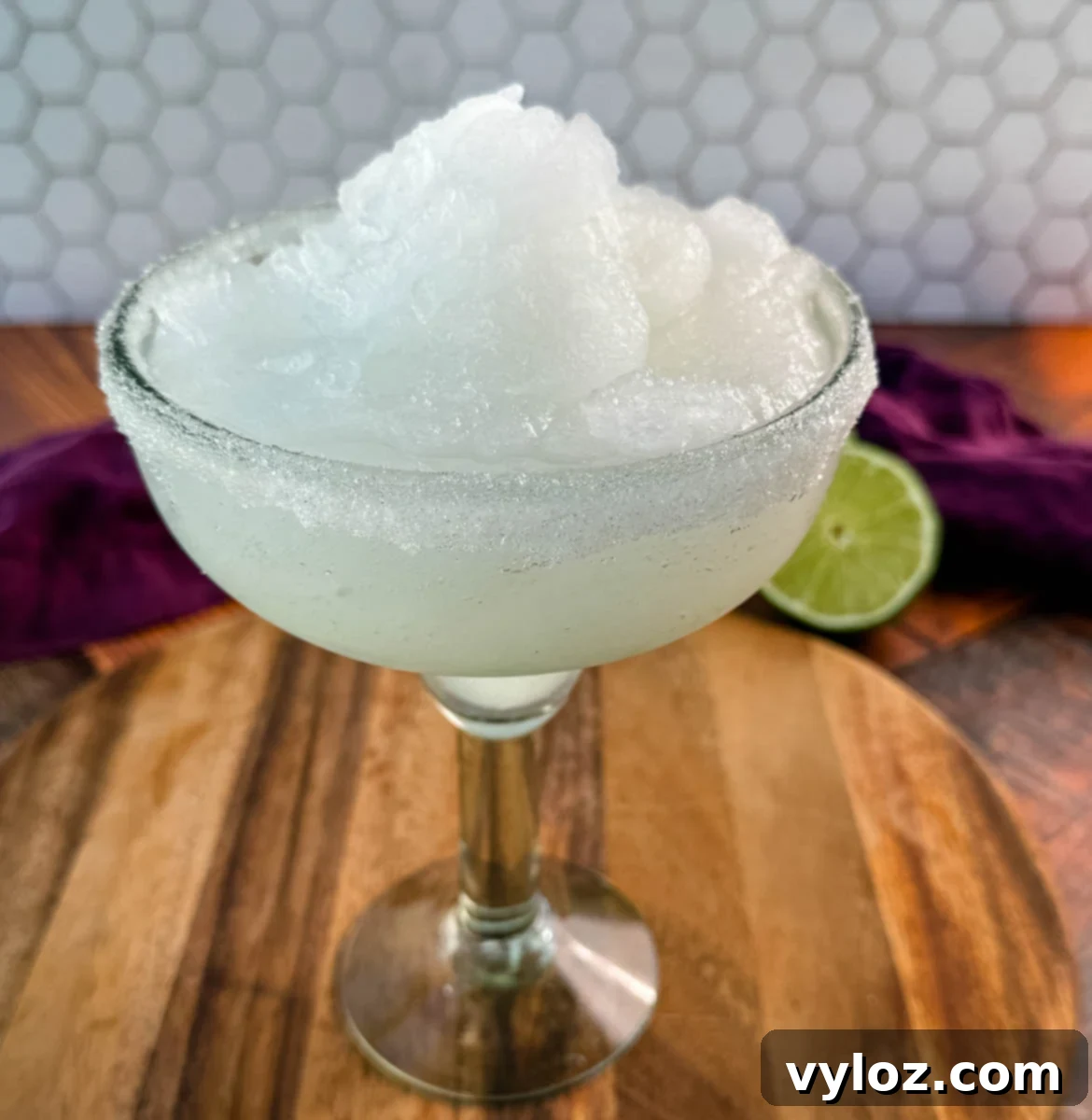 A close-up of a margarita glass filled with frozen slush, highlighting the fluffy, snow-like texture of the drink. The sugar-rimmed glass sits on a rustic wooden board.