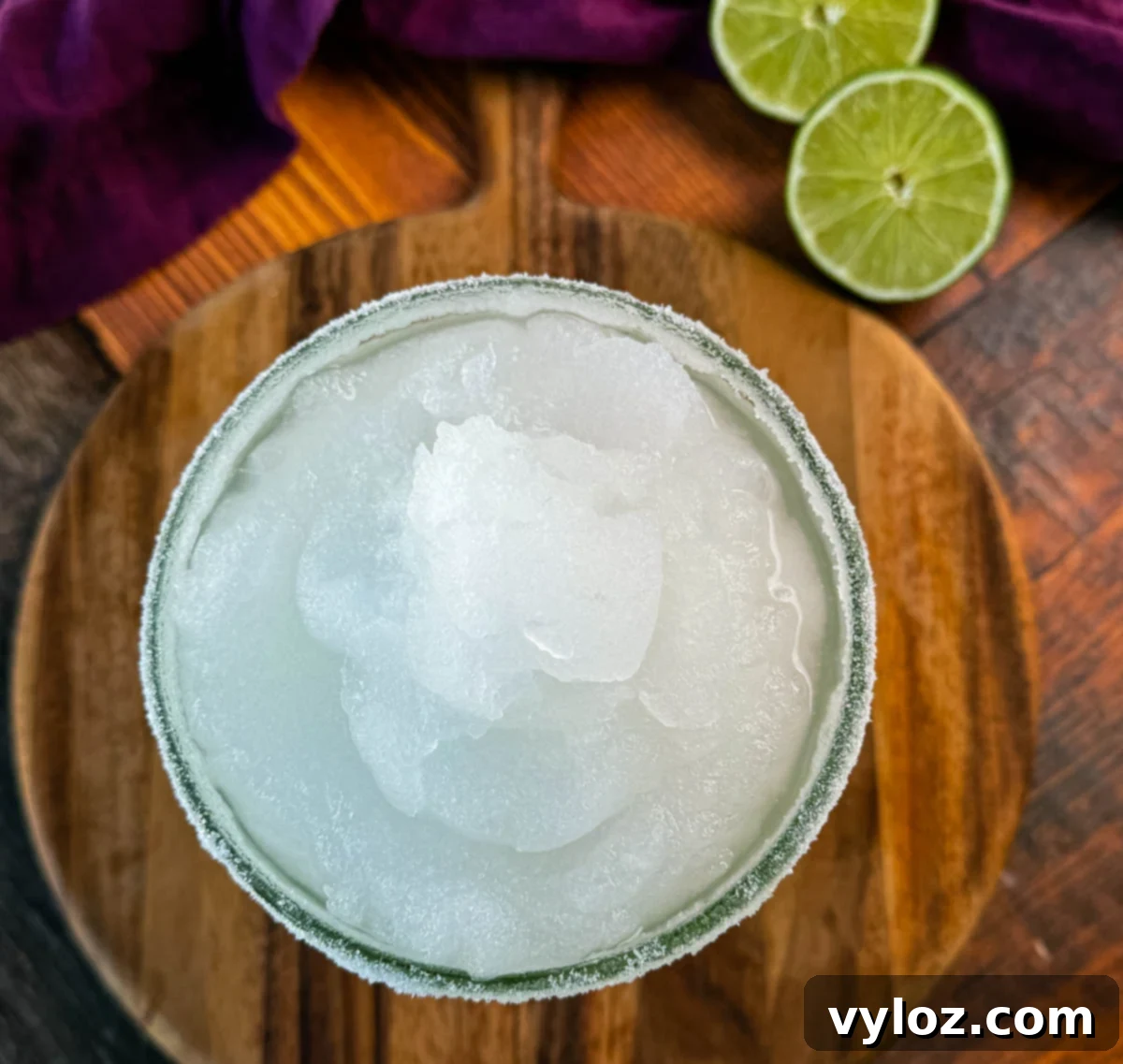 A top-down shot of a frozen margarita in a glass with a sugar rim, showcasing the smooth and icy texture. Fresh lime slices and a purple cloth napkin are visible in the background.