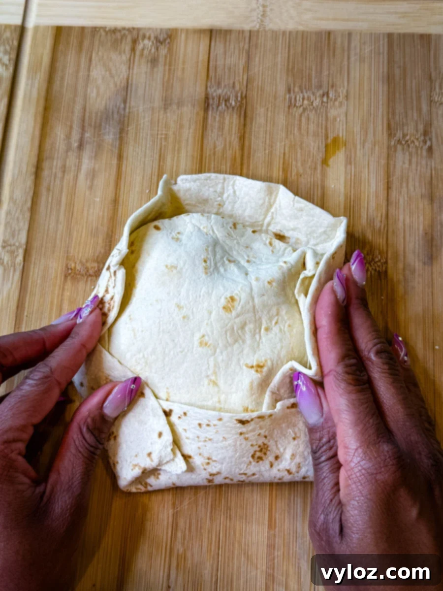 Hands folding the edges of a large flour tortilla over a smaller tortilla-covered filling to create a crunchwrap on a wooden cutting board.