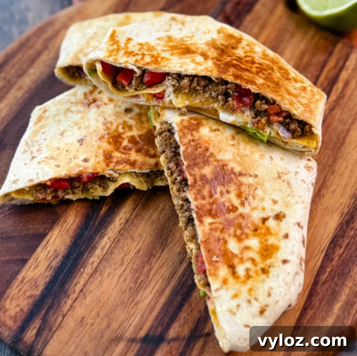 Golden brown beef and cheese crunchwrap supreme cut in half and stacked on a wooden cutting board with halved limes in the background