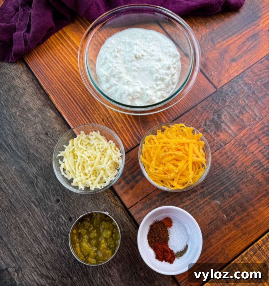 Protein-Packed Queso Dip 3 Flat lay of ingredients for high-protein queso dip on a rustic wood surface. Includes bowls of cottage cheese, shredded white and yellow cheese, diced green chiles, and a small bowl with chili powder, smoked paprika, salt, and pepper.