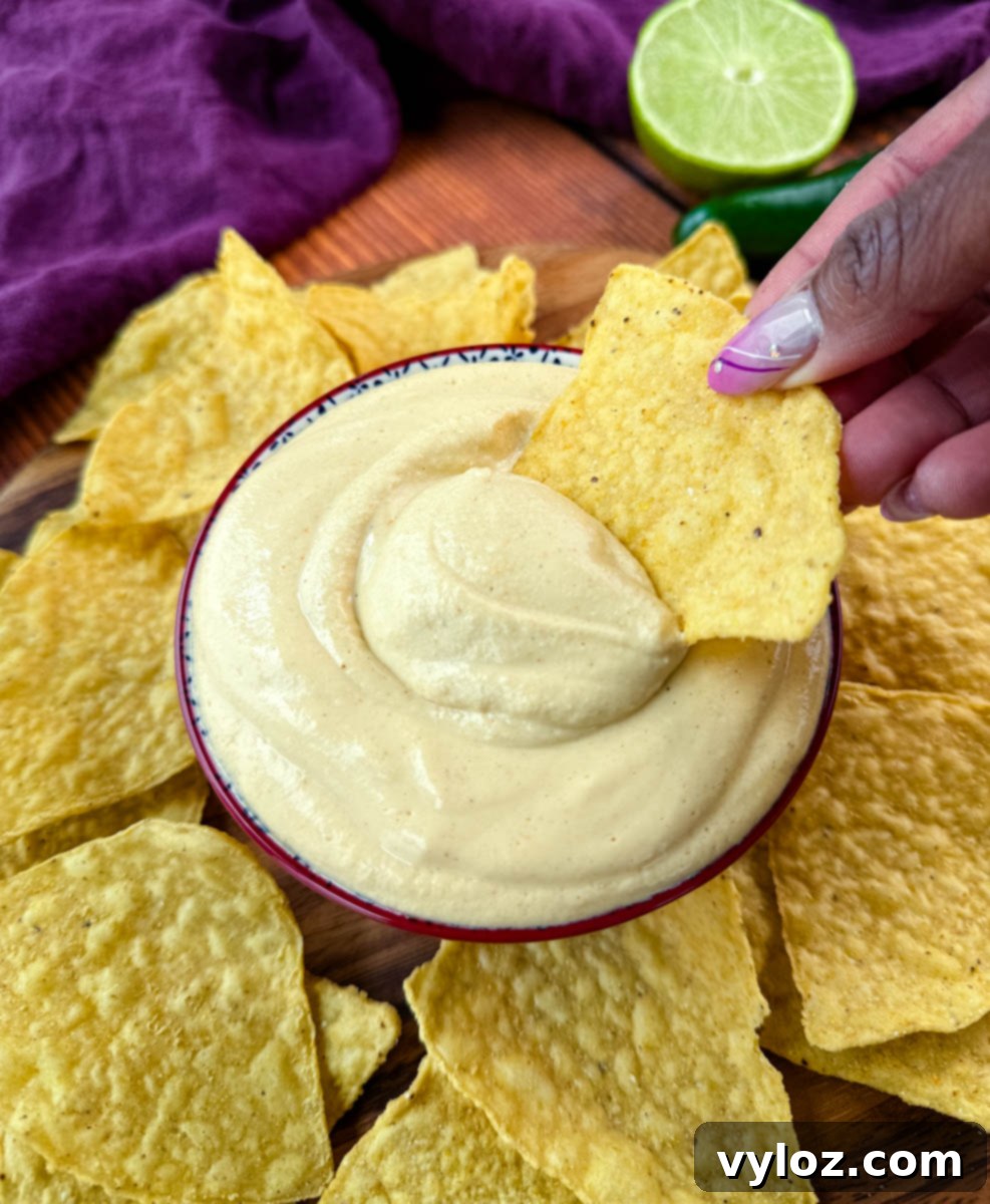 Protein-Packed Queso Dip 8 Hand holding a tortilla chip with swirled cottage cheese queso dip, set against a backdrop of chips, lime halves, and jalapeño slices.