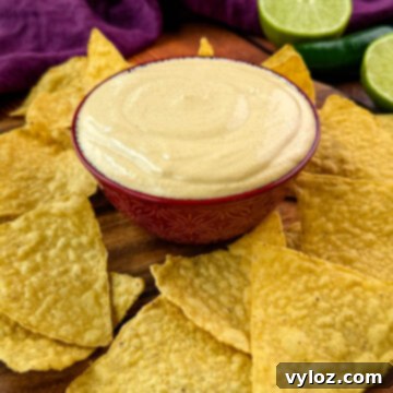 Protein-Packed Queso Dip 10 A decorative bowl filled with creamy high-protein queso dip on a wooden board, surrounded by crispy tortilla chips and garnished with lime and jalapeño.