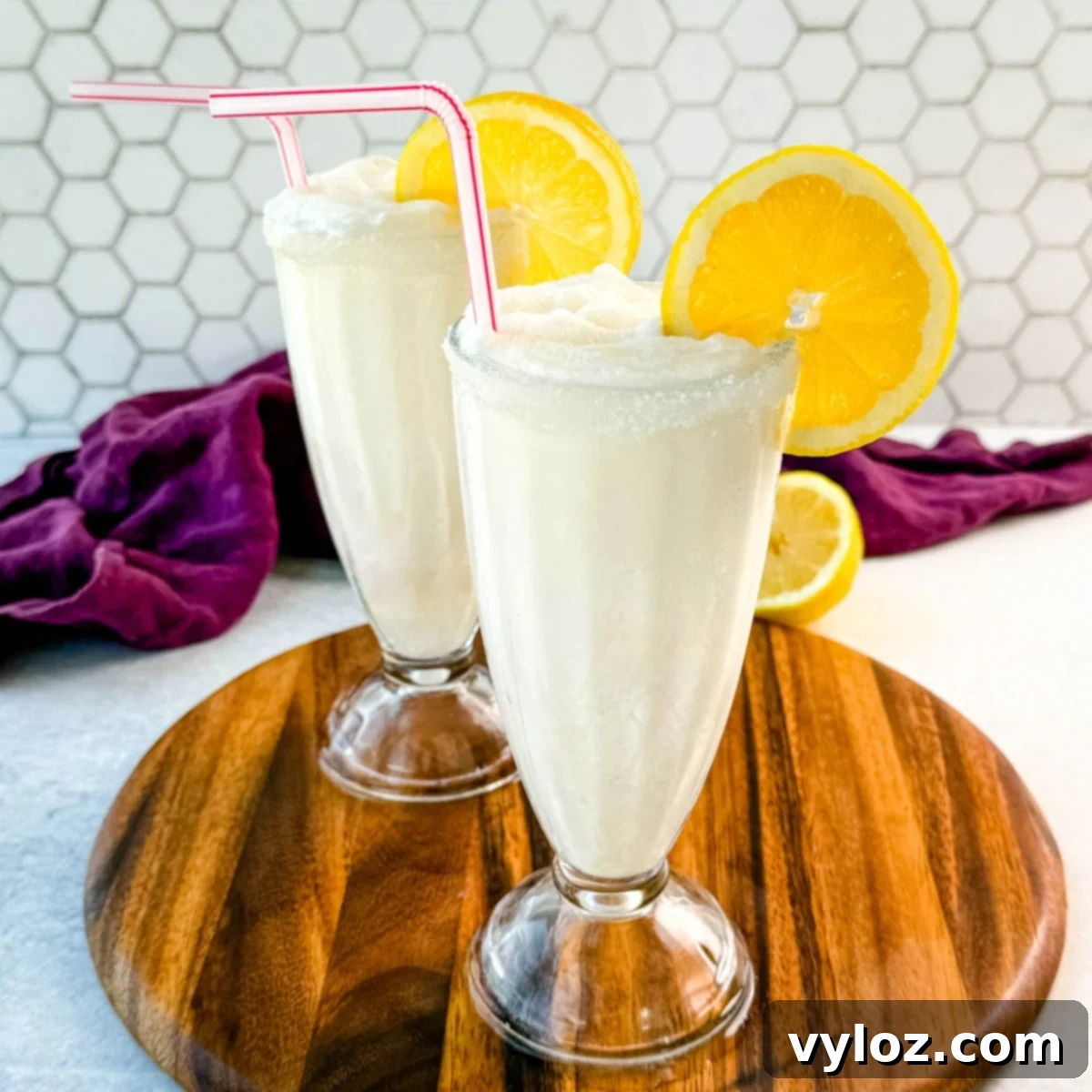 Two finished frosted lemonades in tall glasses, each with a sugared rim, lemon slice, and straw. Placed on a wooden board with a purple napkin and lemon halves in the background.
