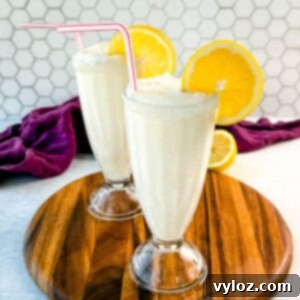 Two finished frosted lemonades in tall glasses, each with a sugared rim, lemon slice, and straw. Placed on a wooden board with a purple napkin and lemon halves in the background.