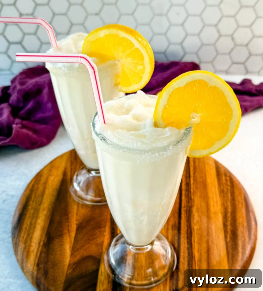 Two finished frosted lemonades in tall glasses, each with a sugared rim, lemon slice, and straw. Placed on a wooden board with a purple napkin and lemon halves in the background.