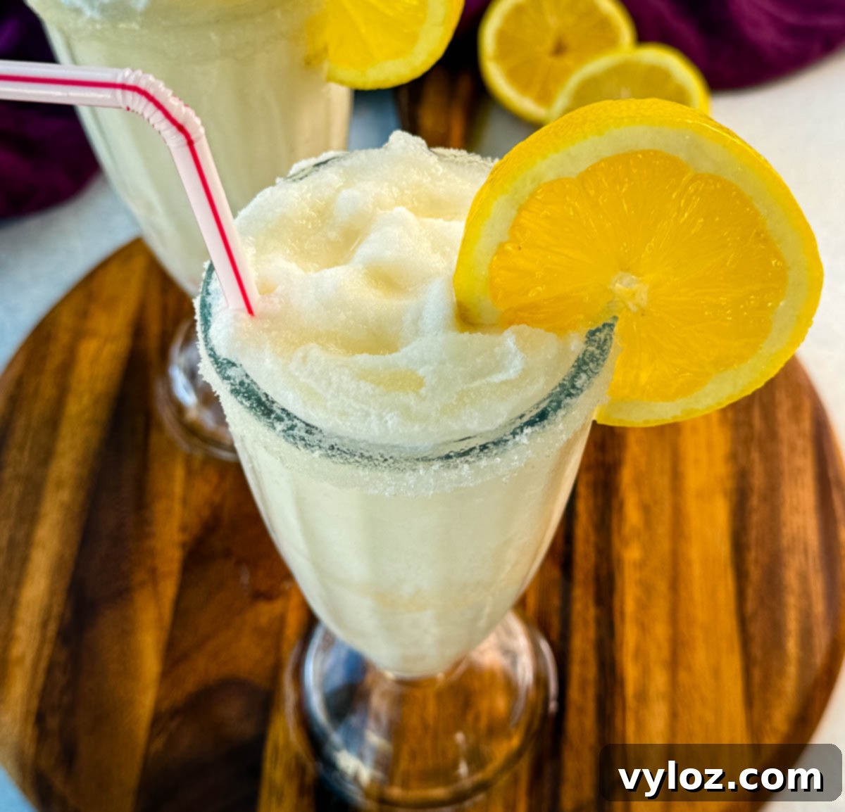 Two finished frosted lemonades in tall glasses, each with a sugared rim, lemon slice, and straw. Placed on a wooden board with a purple napkin and lemon halves in the background.