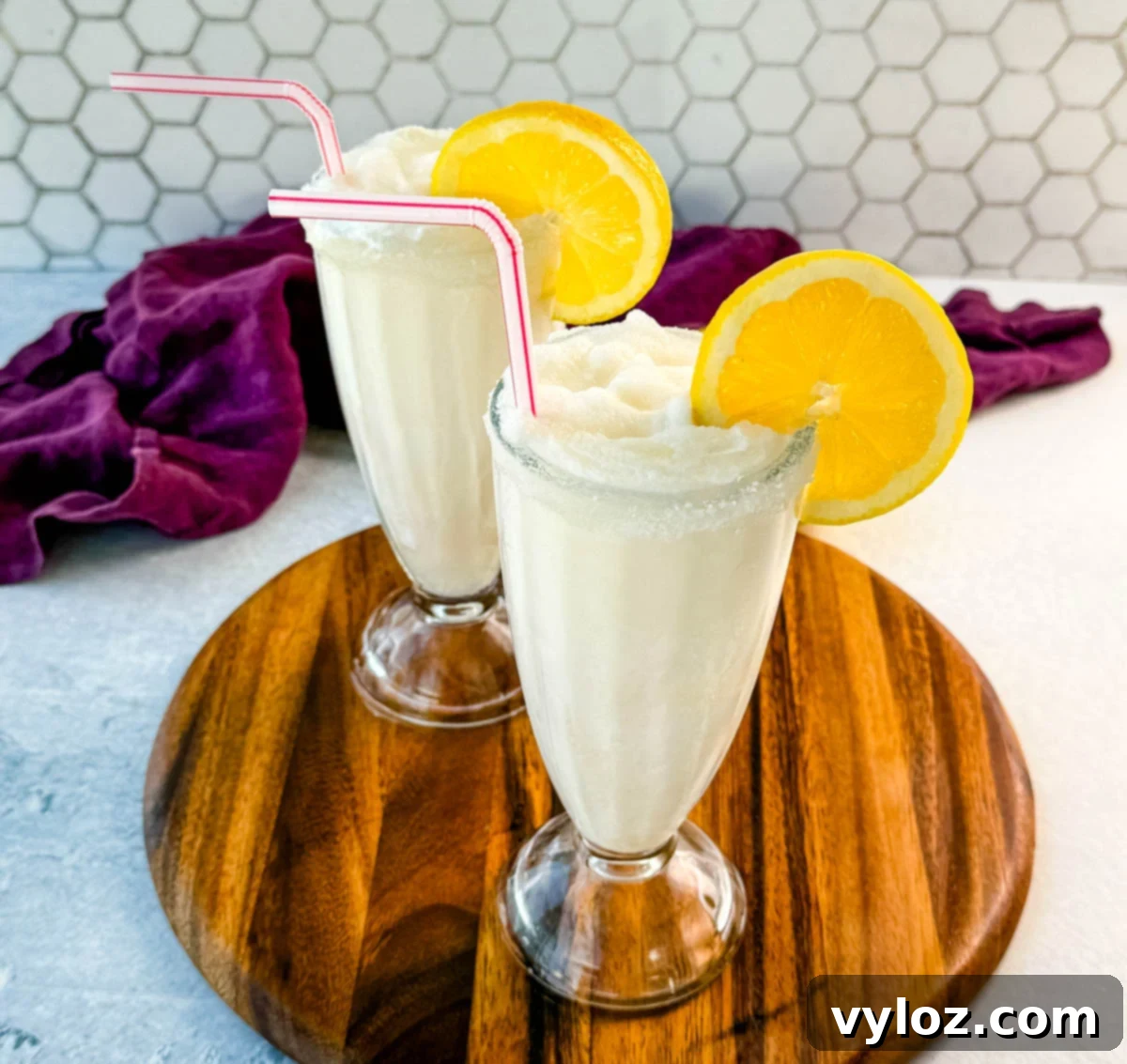 Two finished frosted lemonades in tall glasses, each with a sugared rim, lemon slice, and straw. Placed on a wooden board with a purple napkin and lemon halves in the background.