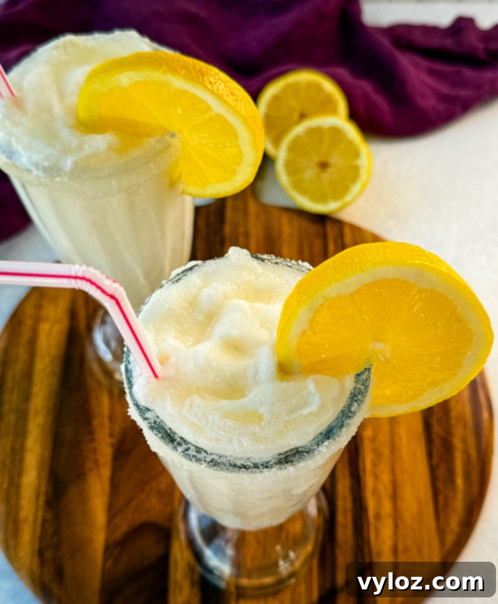 Two finished frosted lemonades in tall glasses, each with a sugared rim, lemon slice, and straw. Placed on a wooden board with a purple napkin and lemon halves in the background.