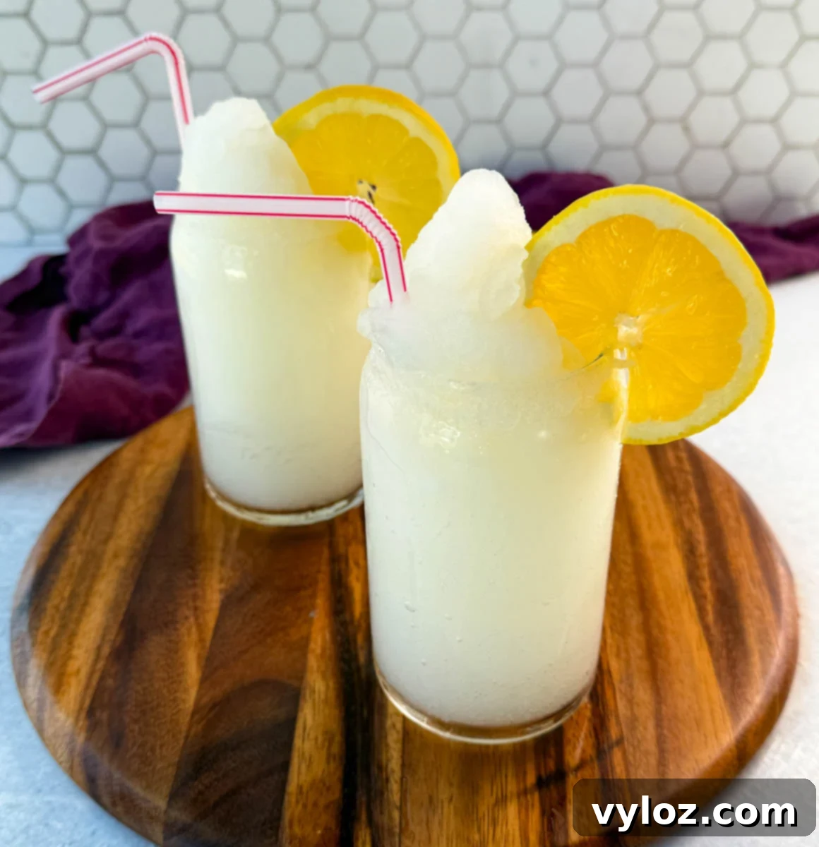 Ninja Lemon Blast 2 Two glasses of lemonade slush on a wooden board, topped with lemon slices and striped straws. A purple cloth and sliced lemons are in the background.