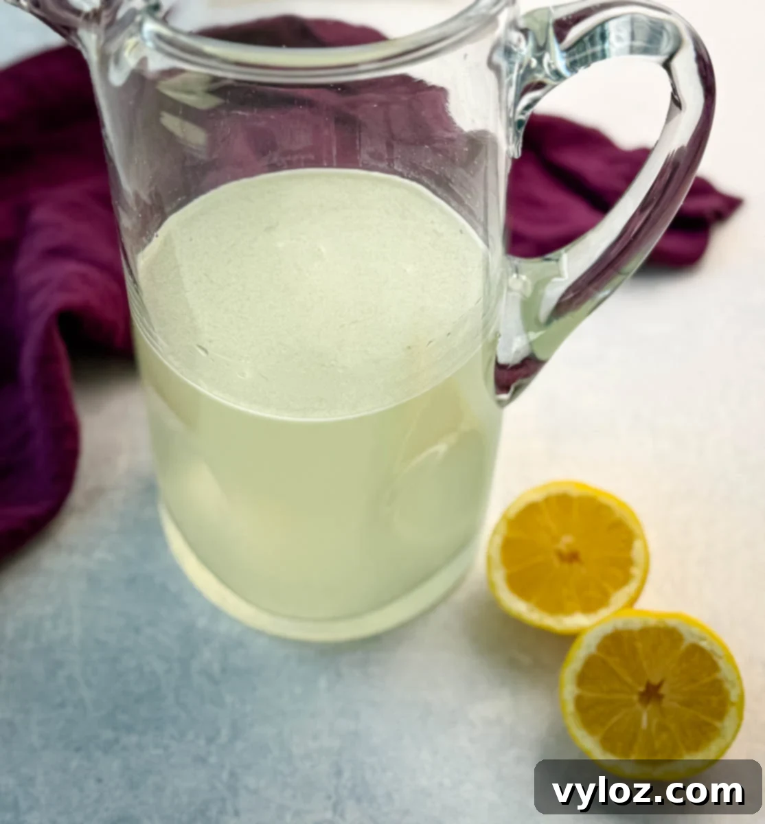 Ninja Lemon Blast 4 A glass pitcher filled with homemade lemonade. Two halved lemons and a deep purple napkin are placed beside it on a light blue surface.