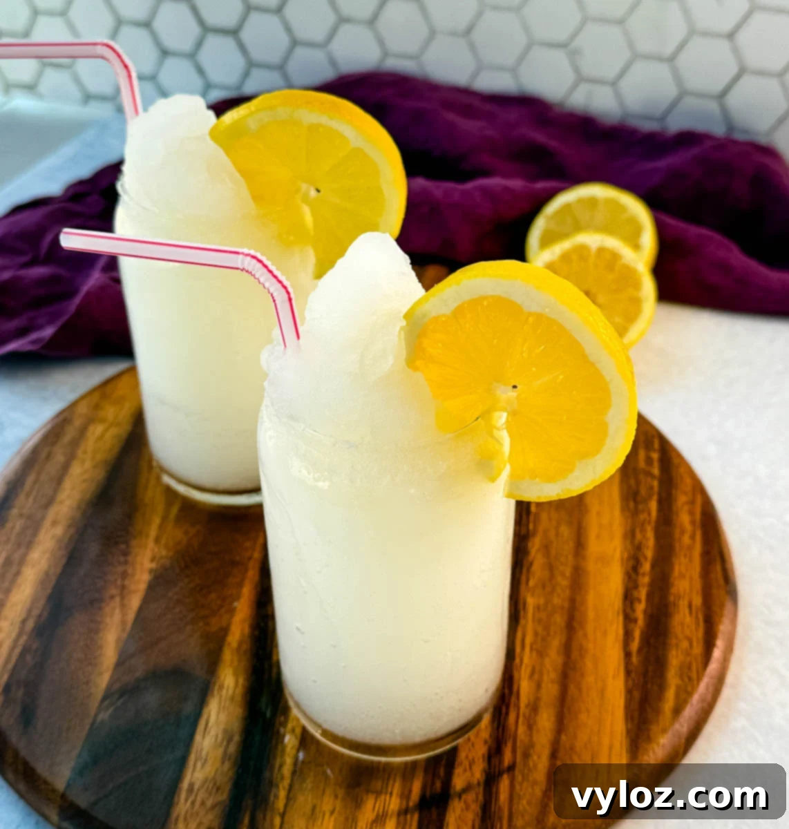 Ninja Lemon Blast 9 Two glasses of lemonade slush on a wooden board, topped with lemon slices and striped straws. A purple cloth and sliced lemons are in the background.