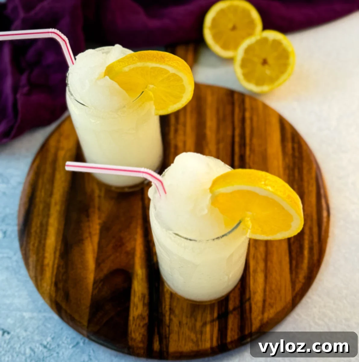 Ninja Lemon Blast 10 Two glasses of lemonade slush on a wooden board, topped with lemon slices and striped straws. A purple cloth and sliced lemons are in the background.