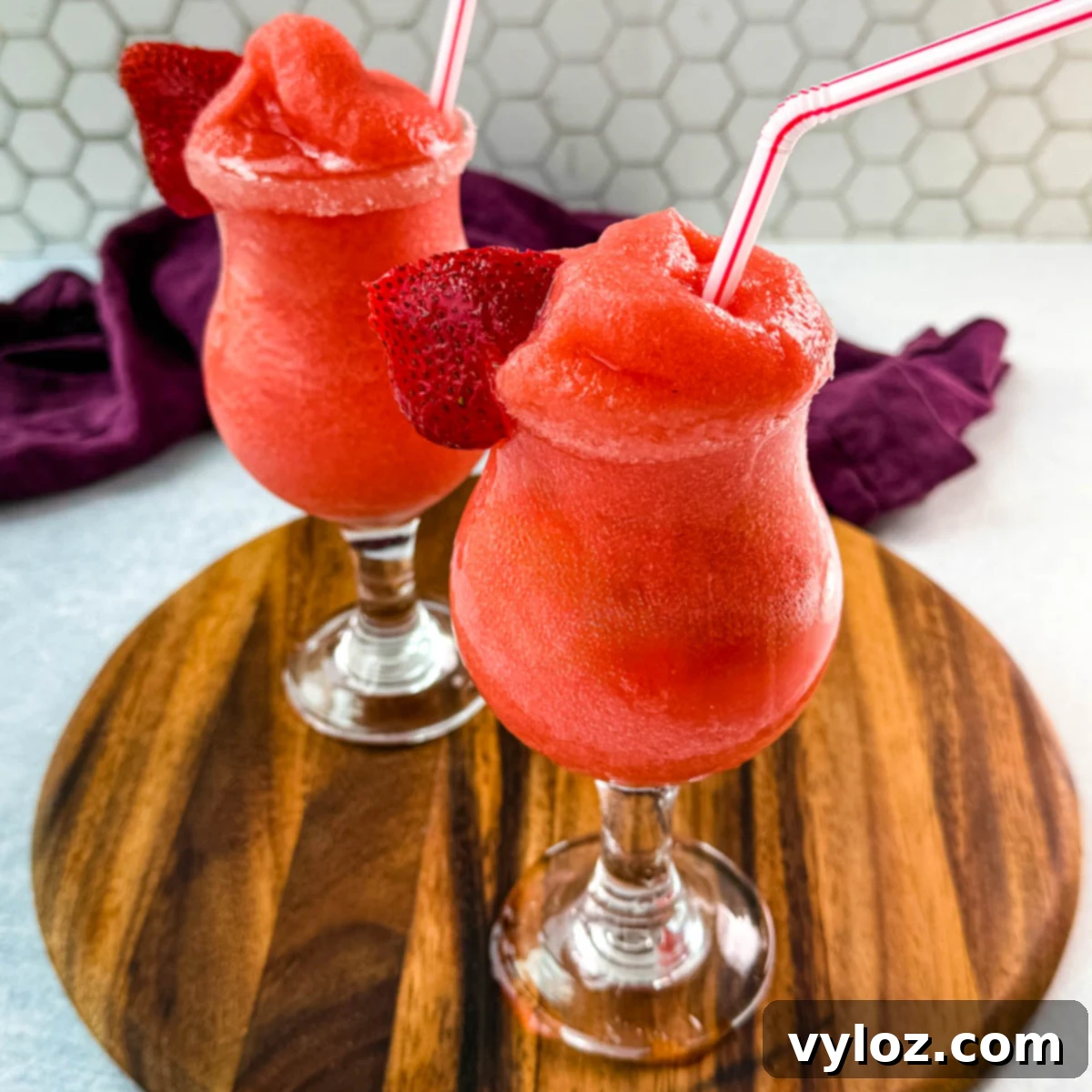 Two glasses of frozen strawberry daiquiris served in curvy cocktail glasses, garnished with fresh strawberry slices and red-striped straws. The vibrant red slushies are placed on a wooden board with a purple cloth in the background.