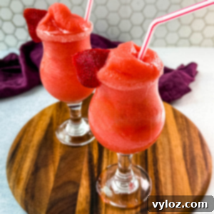 Two glasses of frozen strawberry daiquiris served in curvy cocktail glasses, garnished with fresh strawberry slices and red-striped straws. The vibrant red slushies are placed on a wooden board with a purple cloth in the background.