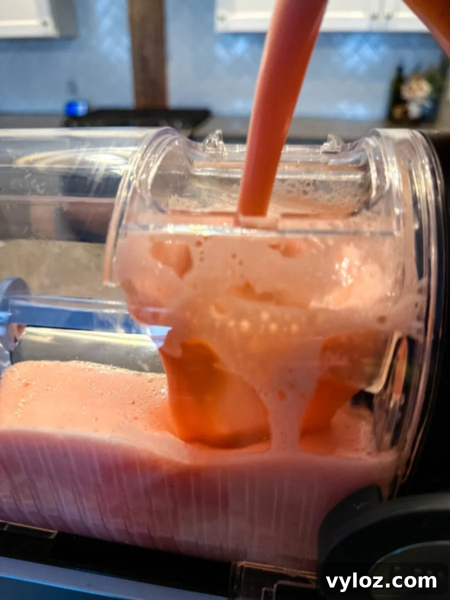 A close-up of a Ninja Slushi machine dispensing a smooth, frozen strawberry daiquiri. The thick pink mixture flows out of the spout into the chamber, highlighting its icy texture.