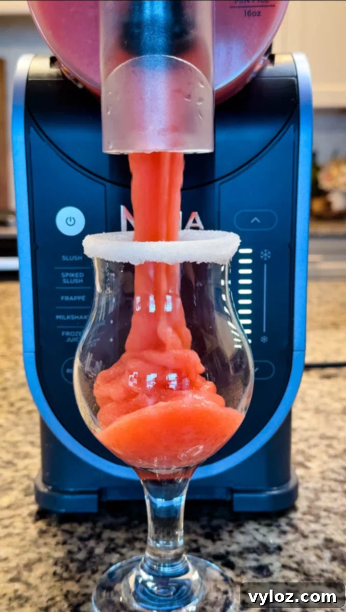 Frozen strawberry daiquiri being dispensed from a Ninja Slushi machine into a sugar-rimmed glass. The bright red slush flows smoothly into the curvy cocktail glass in front of the machine.