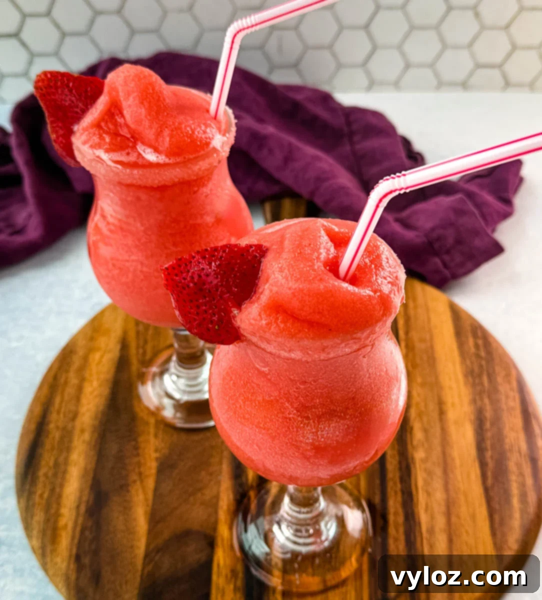 Two glasses of frozen strawberry daiquiris served in curvy cocktail glasses, garnished with fresh strawberry slices and red-striped straws. The vibrant red slushies are placed on a wooden board with a purple cloth in the background.