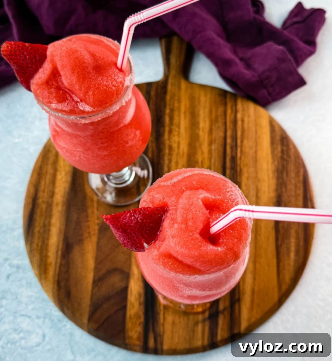 Top-down view of two frozen strawberry daiquiris served in curvy glasses with sugar rims and red-striped straws. Each is garnished with a strawberry slice, placed on a round wooden board with a deep purple napkin in the background.