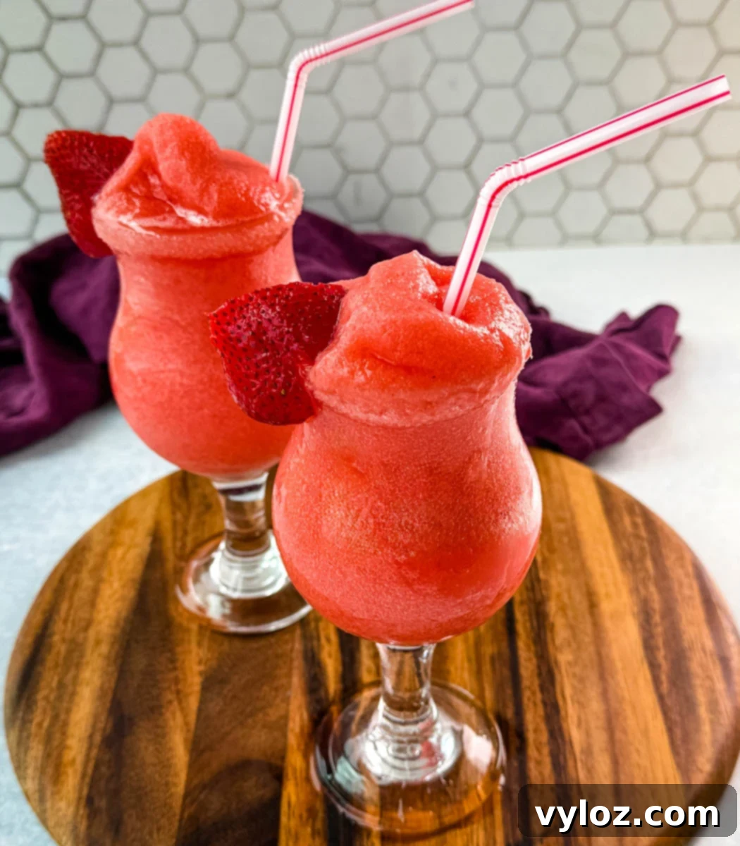 Two glasses of frozen strawberry daiquiris served in curvy cocktail glasses, garnished with fresh strawberry slices and red-striped straws. The vibrant red slushies are placed on a wooden board with a purple cloth in the background.