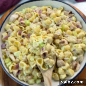 Overhead image of a bowl filled with creamy chicken macaroni salad made with shell pasta, red onion, celery, and chopped grilled chicken. A wooden spoon is scooping some of the salad.