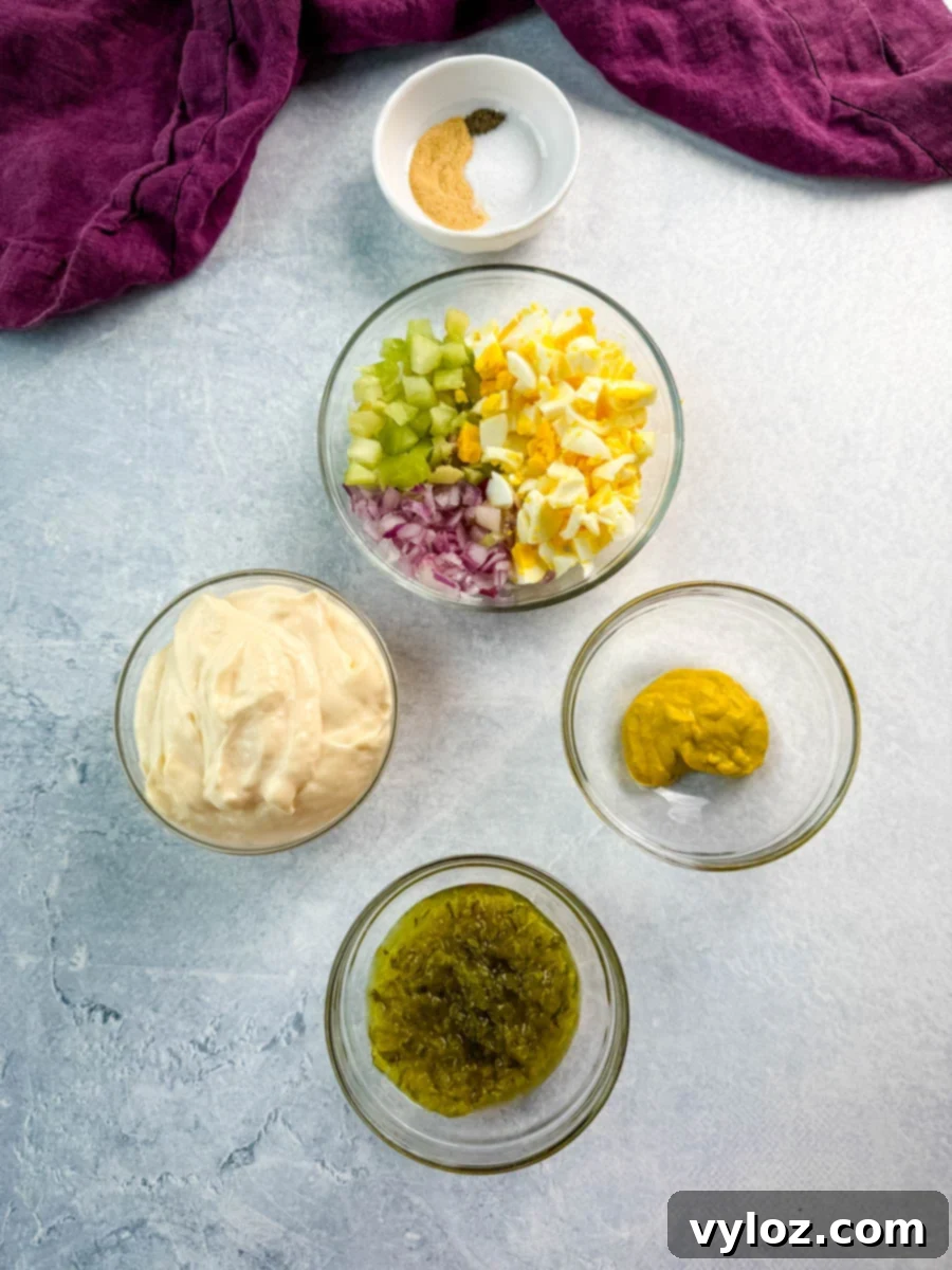 Overhead view of ingredients for chicken macaroni salad arranged in bowls: chopped celery, red onion, and boiled eggs; mayo; mustard; sweet relish; and a small bowl of spices including salt, pepper, garlic powder.
