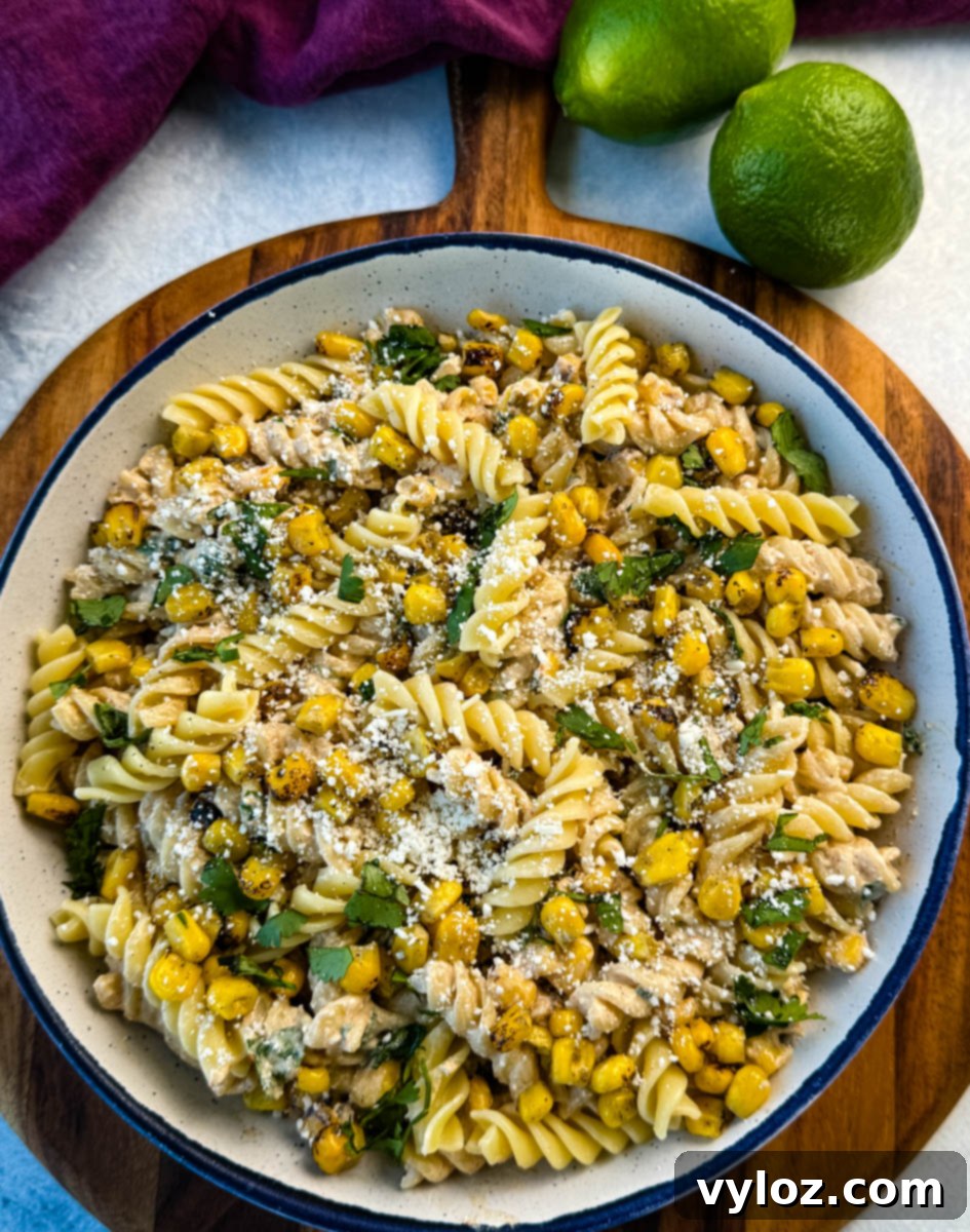 Overhead image of a large bowl of elote pasta salad with rotini, charred corn, crumbled cheese, and chopped cilantro. Two limes sit beside the bowl.