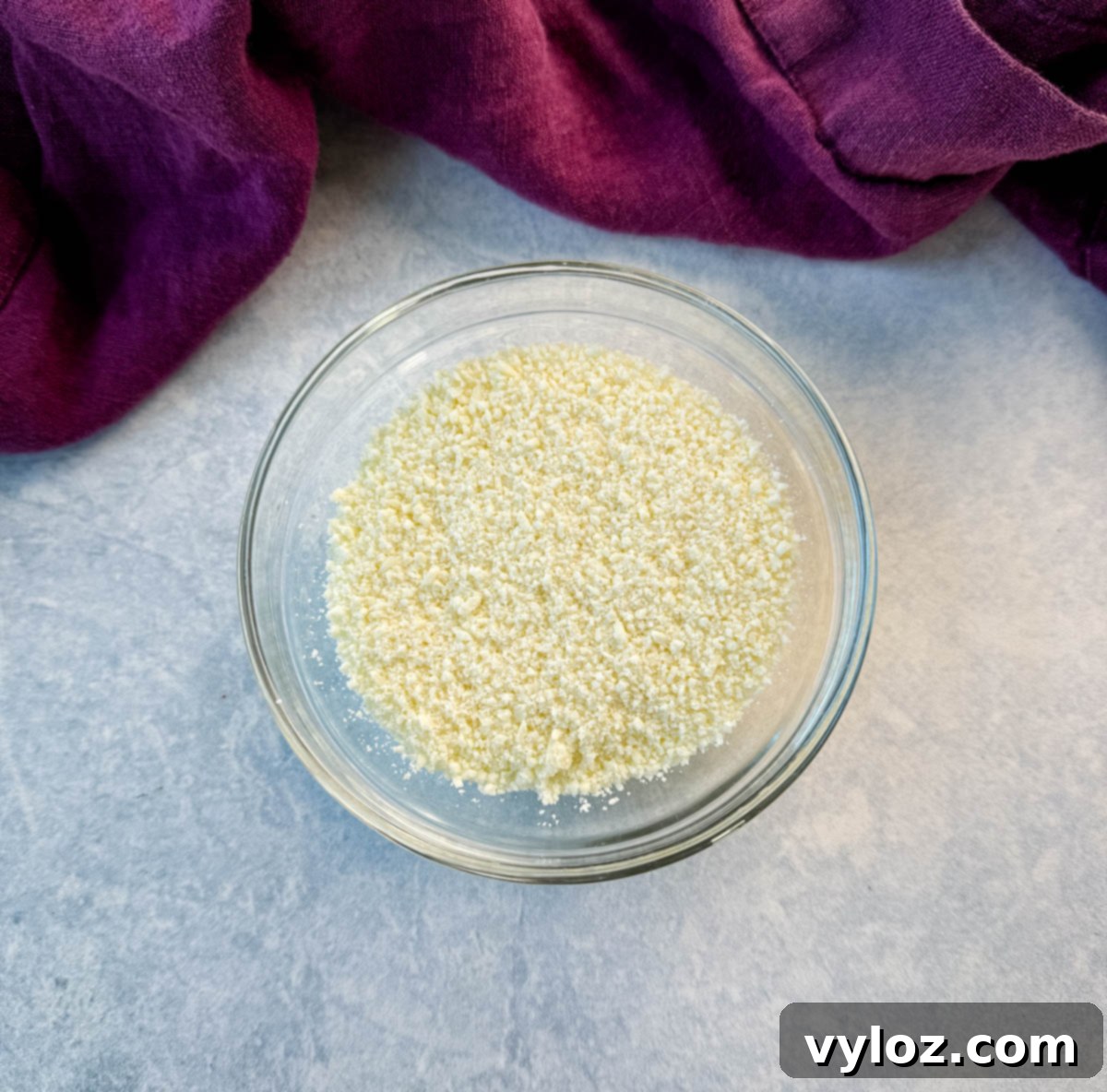 Small glass bowl filled with crumbled cotija cheese, placed on a light countertop with a purple kitchen towel in the background.