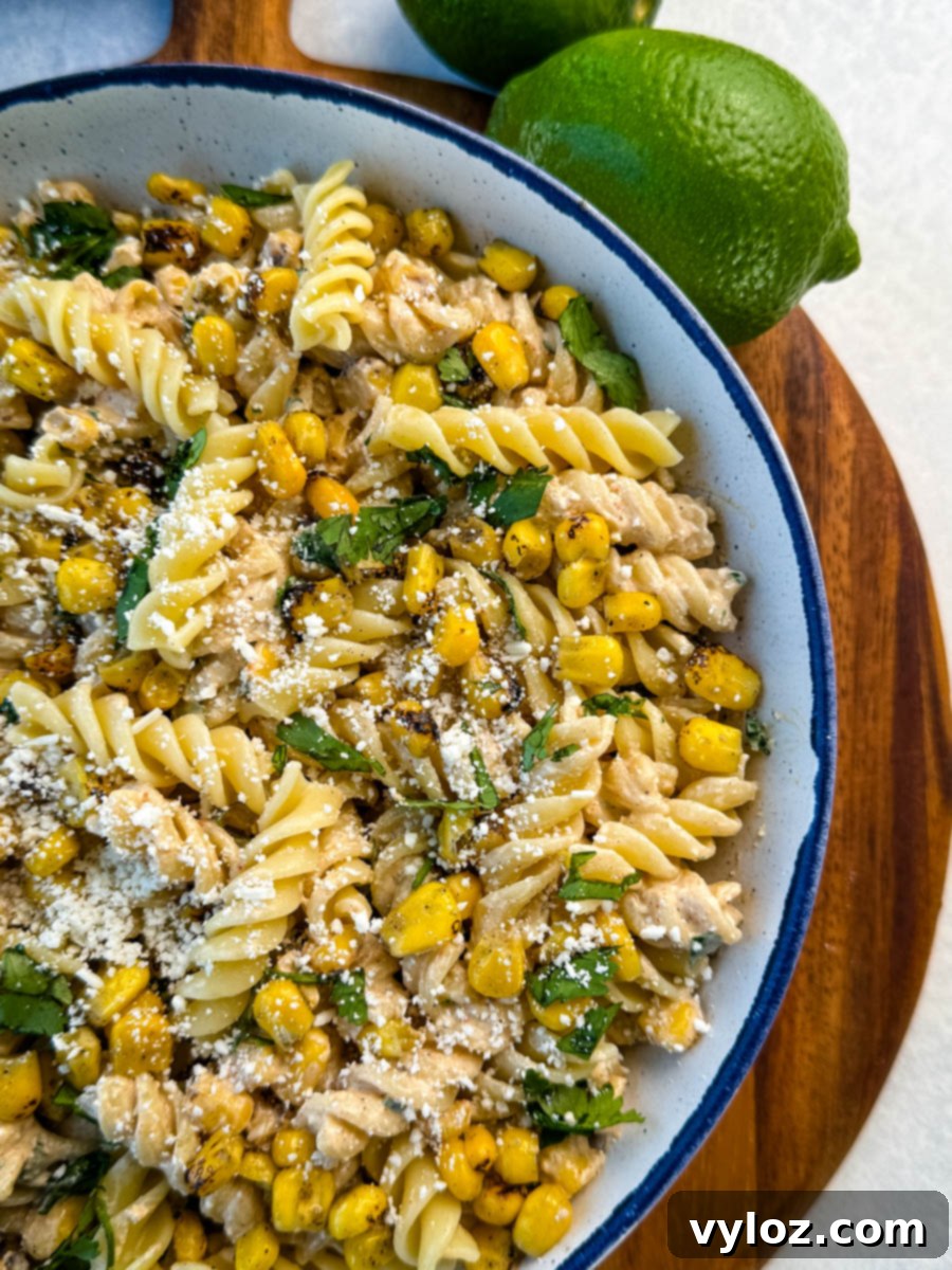 Side view of a serving bowl filled with street corn pasta salad topped with cilantro and cotija cheese, with fresh limes in the background.