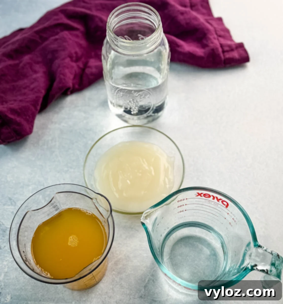 Overhead view of Ninja Slushi piña colada ingredients including pineapple juice, cream of coconut, white rum, and water arranged on a light surface with a purple kitchen towel.