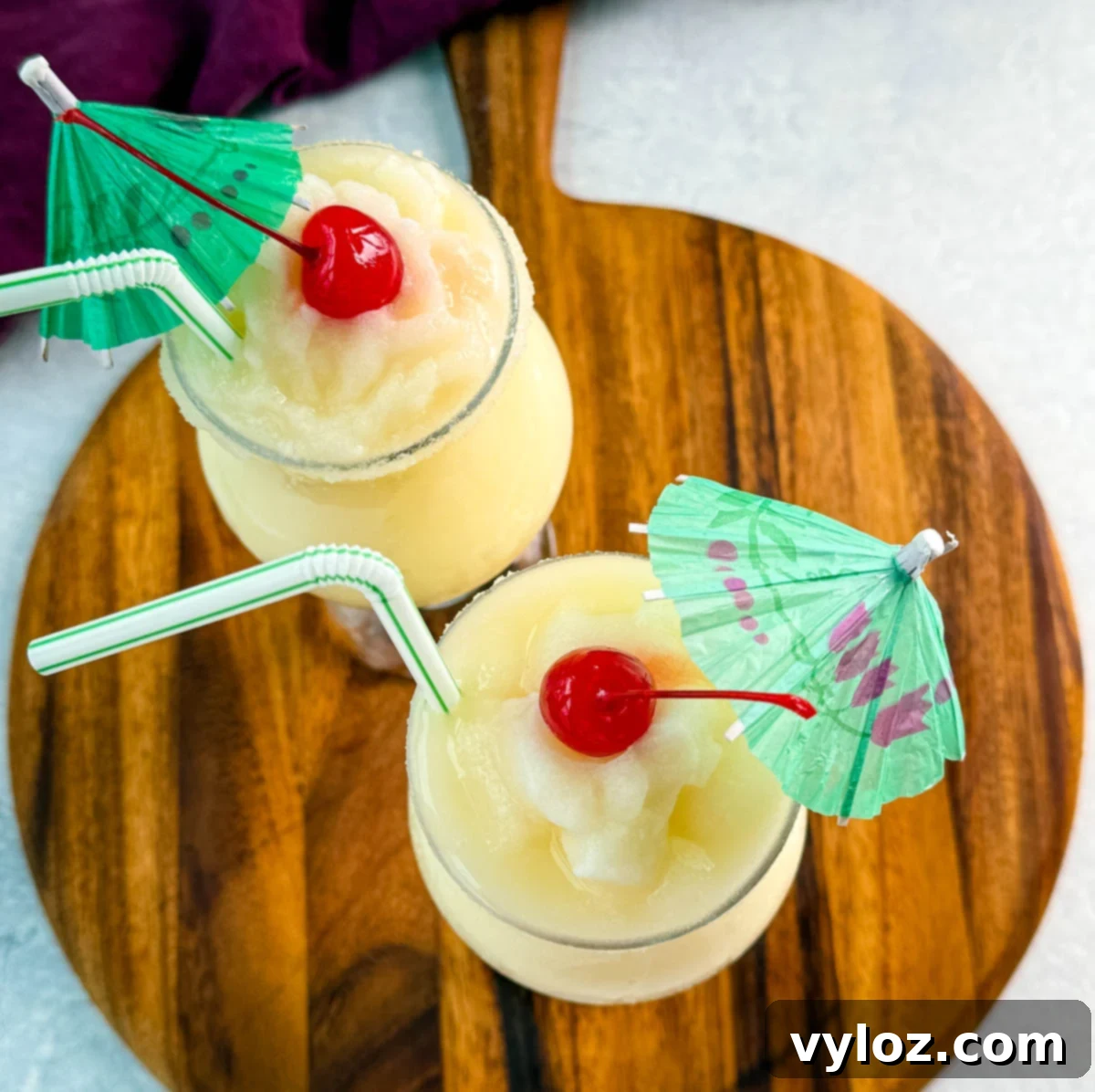 Two frozen piña coladas served in glasses on a wooden board, topped with cherries and paper umbrellas.