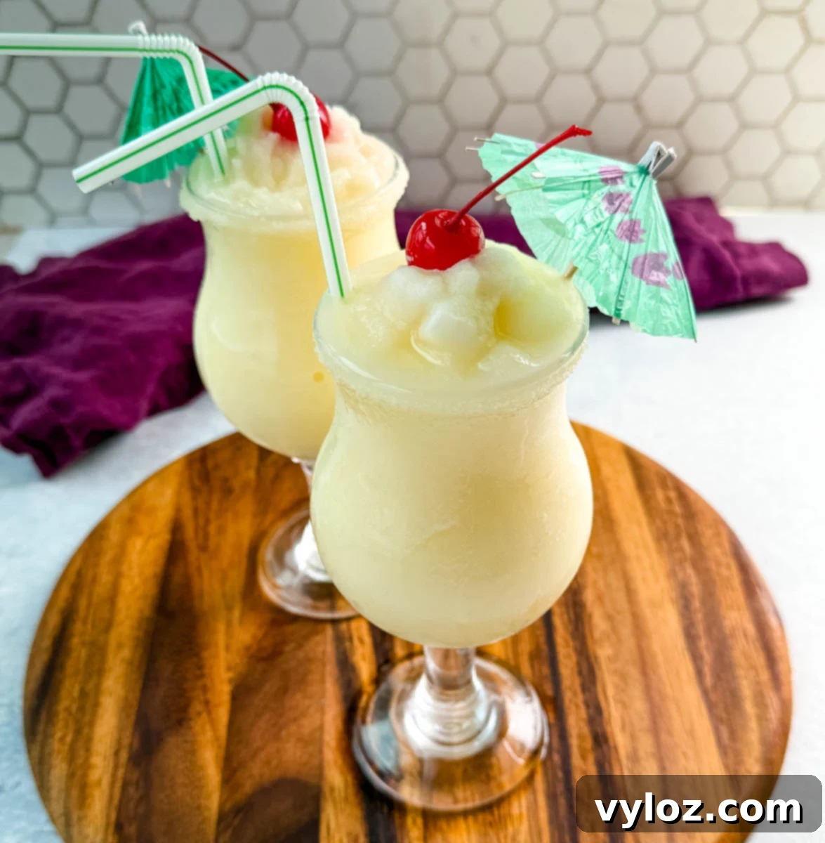 Close-up of creamy piña coladas in curved glasses with straws, cherries, and umbrellas, ready to serve.