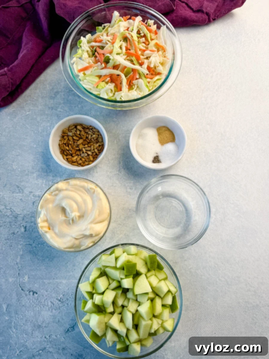 Creamy Apple Crunch Slaw 3 Overhead view of ingredients for creamy apple coleslaw arranged on a light surface, including shredded cabbage and carrots, diced green apples, mayo, seasonings, vinegar, and sunflower seeds.