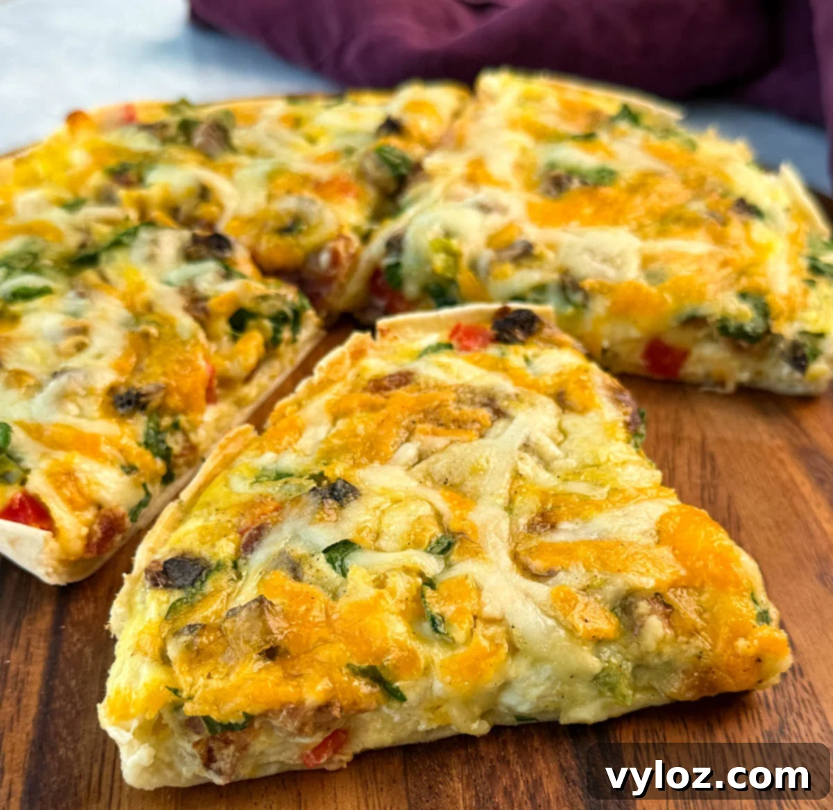 Close-up of a cheesy slice of tortilla egg bake on a wooden board, showcasing the golden melted cheese, bacon, and vibrant vegetables baked into the top.
