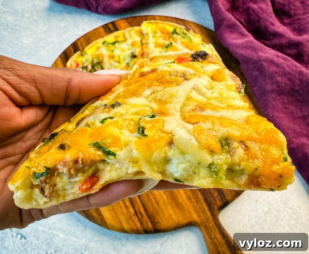 A hand holding a slice of baked egg tortilla pizza loaded with melted cheese, bacon, and colorful vegetables, with the full pizza in the background on a wooden board.