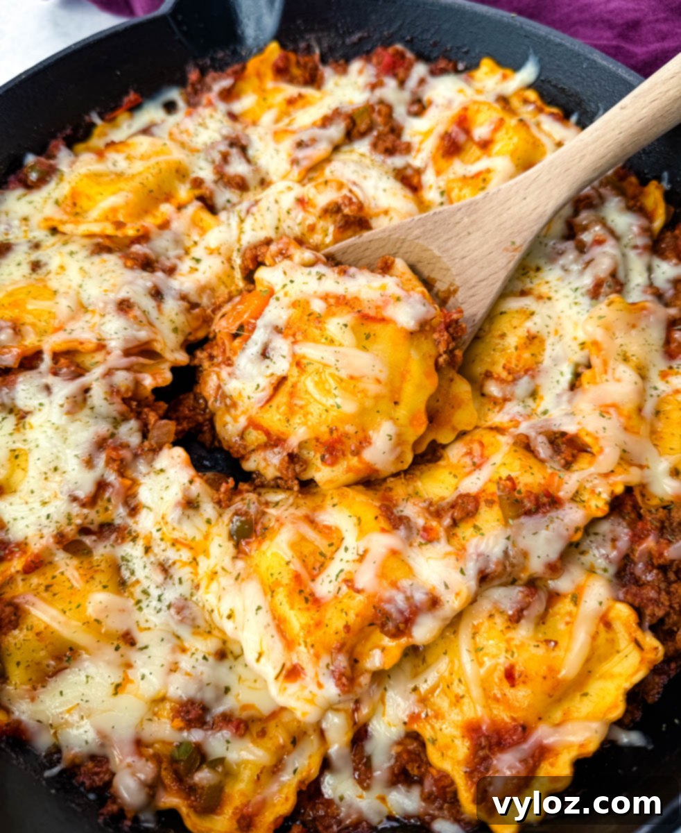 Top-down view of baked ravioli lasagna in a skillet. Ravioli is layered with meat sauce and melted cheese, garnished with dried herbs.