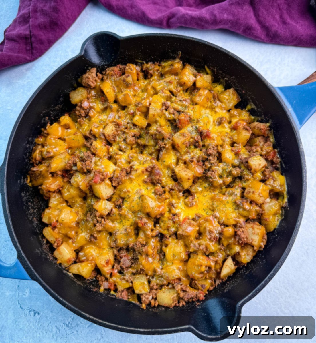 Savory Taco Potatoes 7 Full view of a cast iron skillet packed with cheesy taco potatoes, topped with herbs, sitting on a light blue surface with a purple kitchen towel nearby.