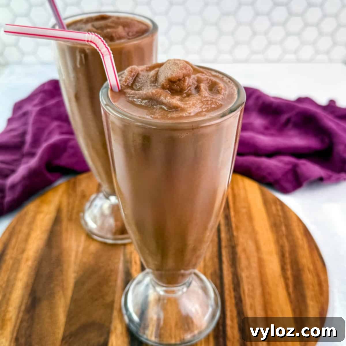 Two chocolate milkshakes served in classic soda fountain glasses on a wood platter, showing off the thick, frozen texture and striped straws.