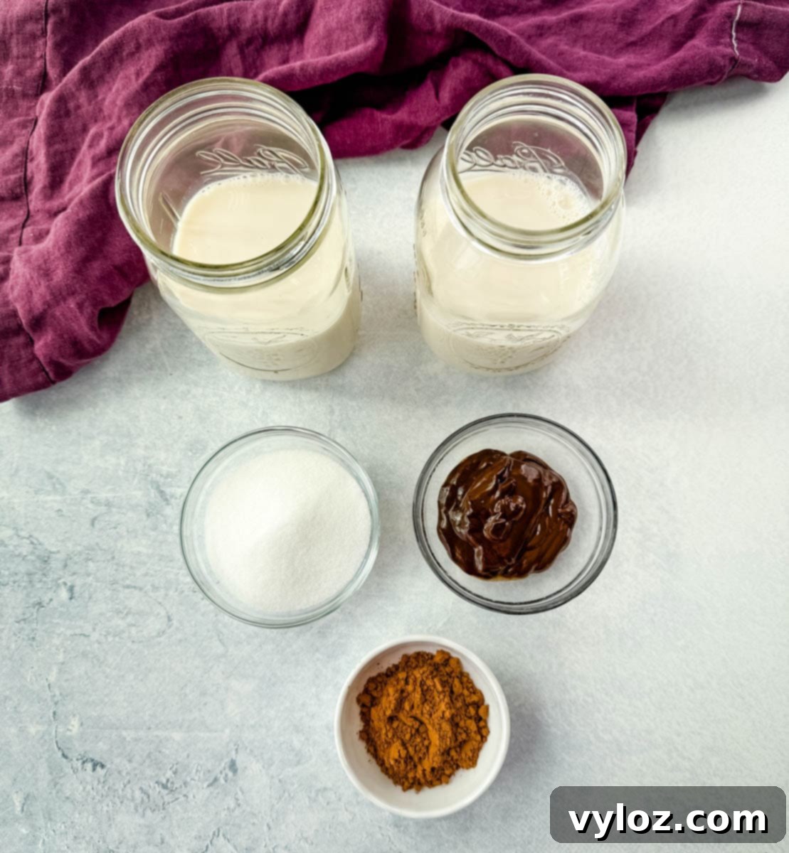 Overhead layout of chocolate milkshake ingredients including two mason jars filled with milk, a small bowl of sugar, a bowl of chocolate syrup, and a dish of cocoa powder, all placed on a light surface with a purple cloth nearby.