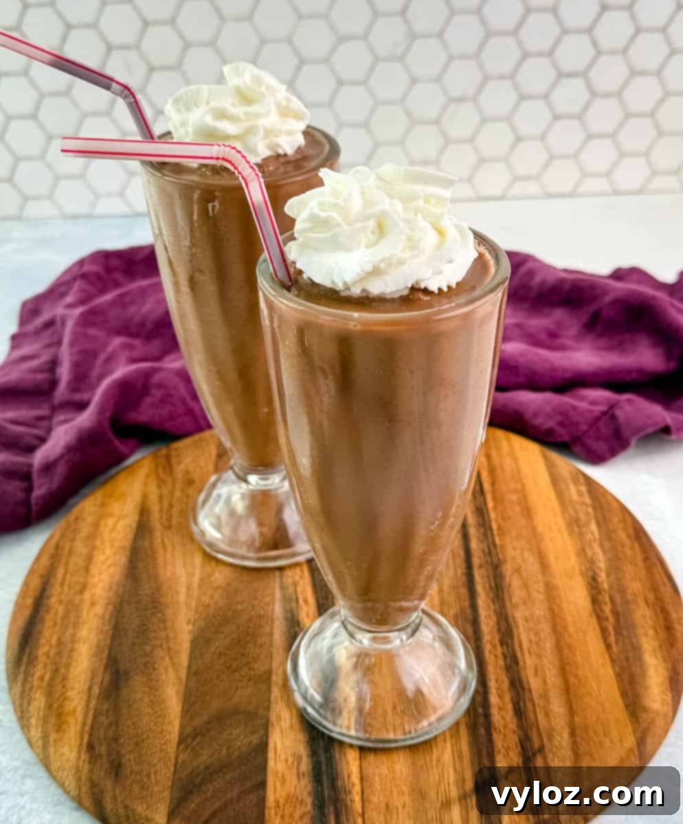 Two tall glass milkshake glasses filled with rich chocolate milkshake, topped with whipped cream and red-striped straws, set on a wooden tray with a purple cloth in the background.