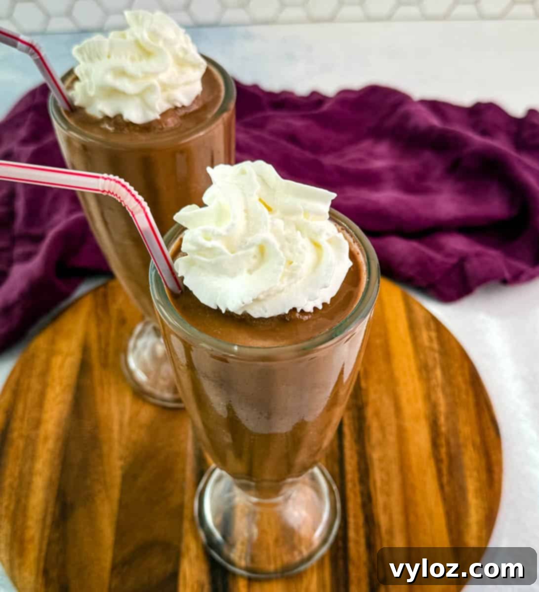 Top-down view of two chocolate milkshakes with whipped cream and red-striped straws, sitting on a round wooden board with a purple napkin in the background.